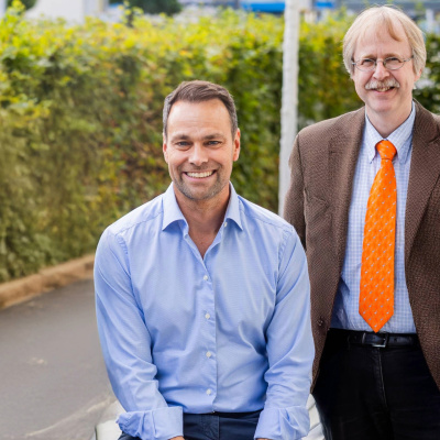 Daniel Fischer sitzt auf einer Bank. Alexander Bracht steht neben ihm. Dahinter ist eine Hecke.
