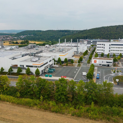 Fabrikgelände, das in einer Landschaft auf Felder und Wald liegt. Im Hintergrund ist ein Dorf.