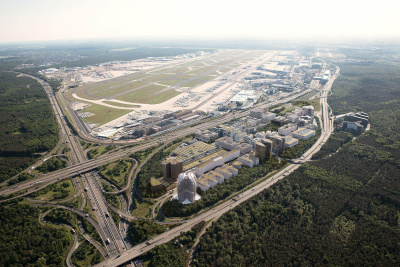 Auf dem Bild ist eine großflächige Luftbild-Visualisierung des Frankfurter Flughafens und des angrenzenden Business-Quartiers Gateway Gardens zu sehen. Das dicht bebaute Areal im Zentrum zeigt das moderne Airport-Quartier mit zahlreichen Bürokomplexen, Hotels und Grünflächen. Markant im unteren Bereich sticht das wellenförmige bzw. geschwungene Hochhausgebäude hervor.