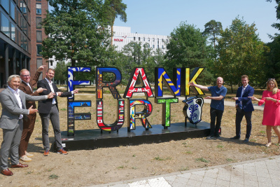 Auf dem Bild ist eine Gruppe von sechs Personen zu sehen, die für ein Foto neben dem mobilen „FRANKFURT“ Selfie-Point posieren. Die Installation besteht aus großen, dreidimensionalen Buchstaben, die den Stadtnamen bilden, ergänzt durch das traditionelle Frankfurter Symbol eines blau-weißen Apfelwein-Bembels am rechten Rand. Jeder Buchstabe ist individuell und farbenfroh mit verschiedenen Mustern wie Flaggen, Wahrzeichen oder dem Frankfurter Adler gestaltet. Im Hintergrund befindet sich das moderne, weiße Gebäude des HOLM (House of Logistics and Mobility), welches im Frankfurter Stadtteil Gateway Gardens liegt.