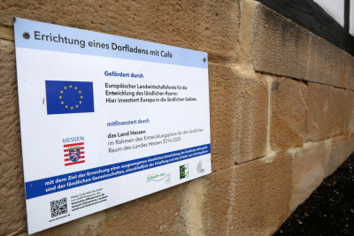 Ein Schild an einem Haus, das darauf hinweist, dass das Land Hessen die Errichtung eines Dorfladens mit Café gefördert hat.