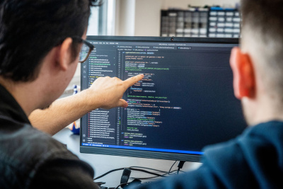 Zwei Männer vor einem Computermonitor auf dem Code dargestellt wird.