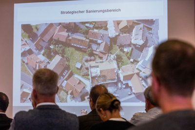 Auf einer Leinwand sind mehrere Häuser zu sehen und der Text "Strategischer Sanierungsplan".