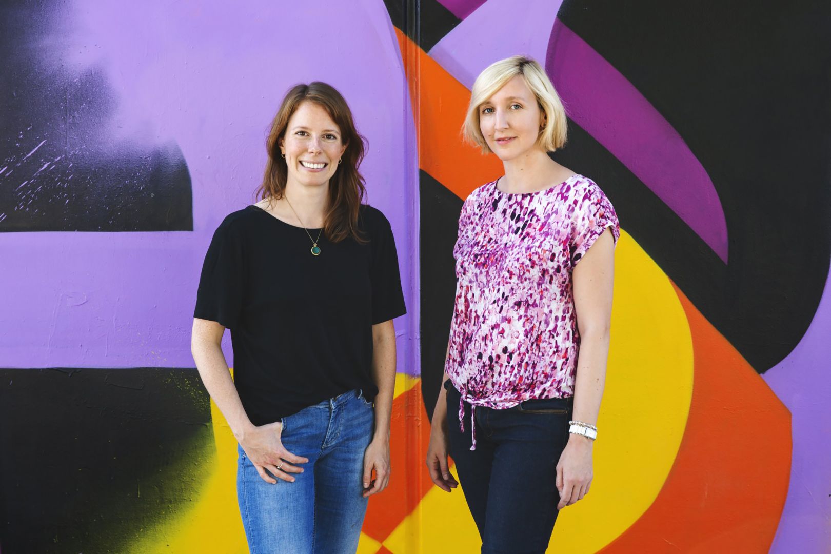 Das Team der Geschäftsstelle Kreativwirtschaft Hessen: Daniela Hartmann und Susanne Stöck stehen vor einer bunten Wand.